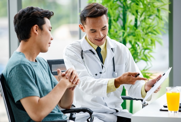 Asian medical practitioner pointing at clipboard and explaining prescription to man during work in hospital