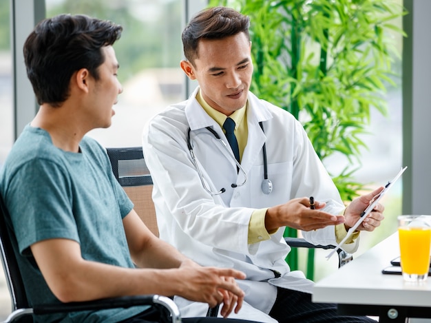 Asian medical practitioner pointing at clipboard and explaining prescription to man during work in hospital