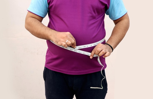 Азиатский мужчина меряет свой толстый живот сантиметровой лентой на однотонном фоне