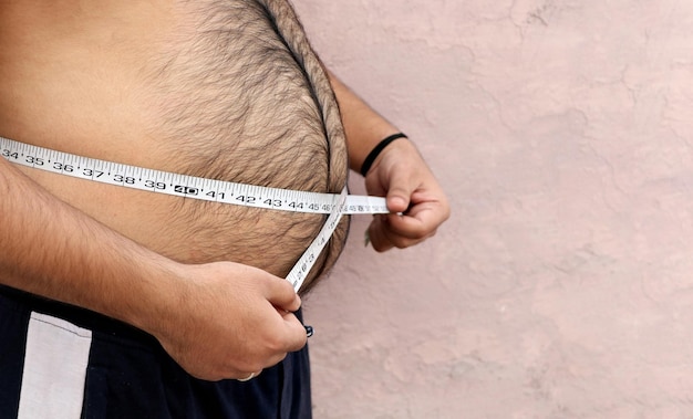 A asian men measures his fat belly with a measuring tape on a plain background