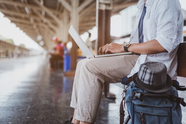아시아 남성들은 여행하기 위해 기차를 타기 위해 기다리는 동안 노트북을 사용합니다. 휴가, 여행, 여행, 여름 여행 컨셉입니다.