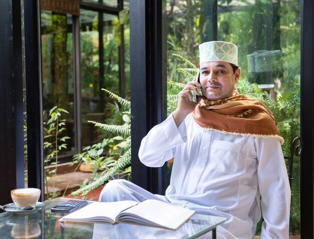 Азиатский средний постаретый мусульманский бизнесмен сидит в кофейне питья кофе с умным мобильным телефоном на таблице.