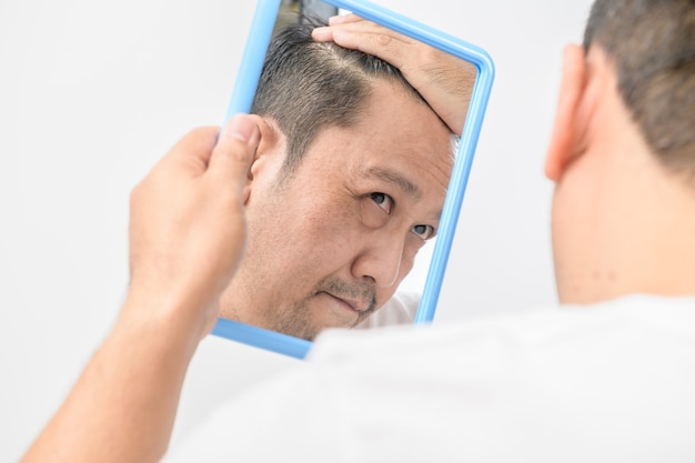 Азиатский мужчина средних лет смотрел в зеркало и беспокоился о выпадении волос или о том, что волосы седые, изолированы на белом фоне, концепция здравоохранения