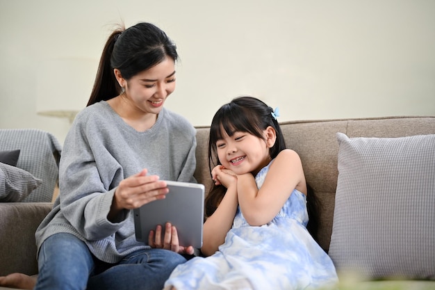 Asian mom showing something on tablet to her daughter watching\
kid\'s cartoon together