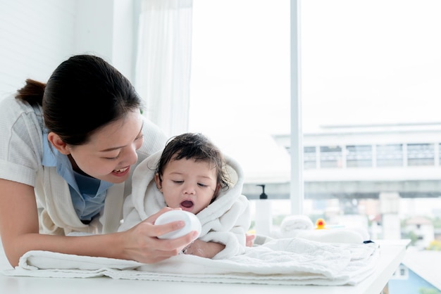 La madre asiatica attraente e giovane sta applicando il borotalco a sua figlia di 7 mesi dopo il bagno