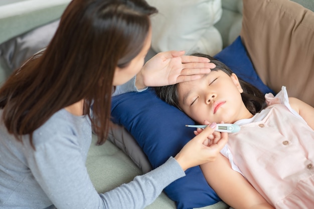 Азиатская мать проверяет температуру тела своего ребенка с помощью термометра, у которого дома жар и болезнь.