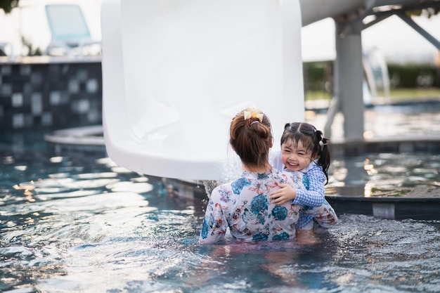 Asian mother and daughter swimming playing slide pool in the\
pool at the resort smiling and laughing having fun in the pool at\
the resort hotel family happy concept