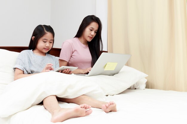 Asian mother and daugther looking laptop and tablet on bed in bedroom