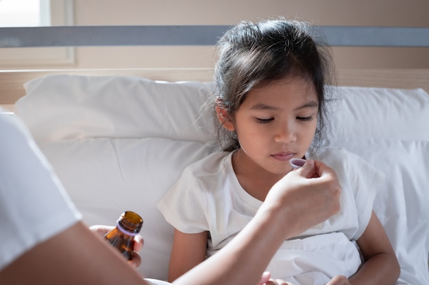 Asian mother giving cough syrup medicine on a spoon to her daughter in bed