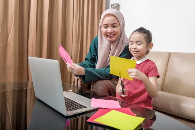 Asian mother helping little girl doing her homework with laptop at home. online education during quarantine