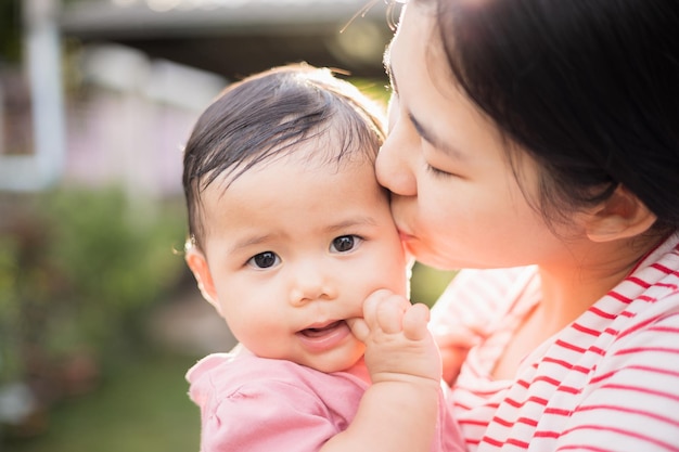 Asian mother holding daughter in arms She held her daughter in her embrace Parents embracing kissing little baby outdoor with sunny day