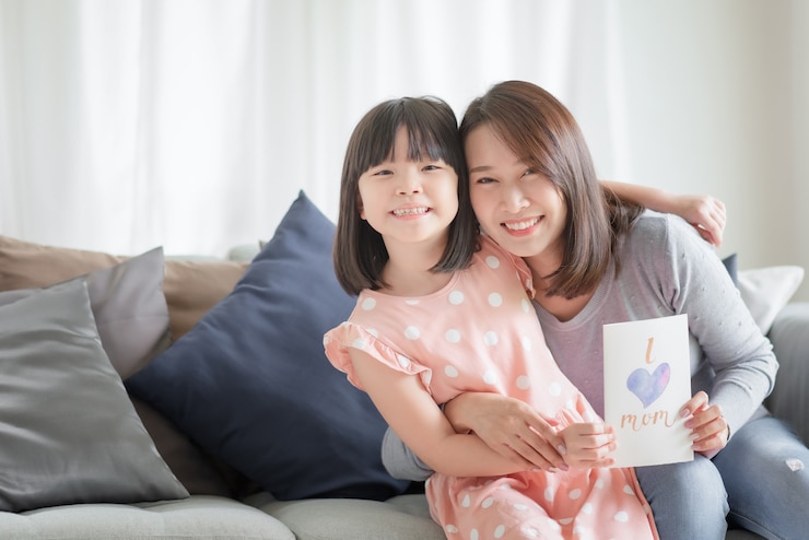아시아 어머니는 집에서 그녀를 놀라게하기 위해 엄마 단어를 사랑하는 수제 인사말 카드를주는 귀여운 딸을 안아줍니다.