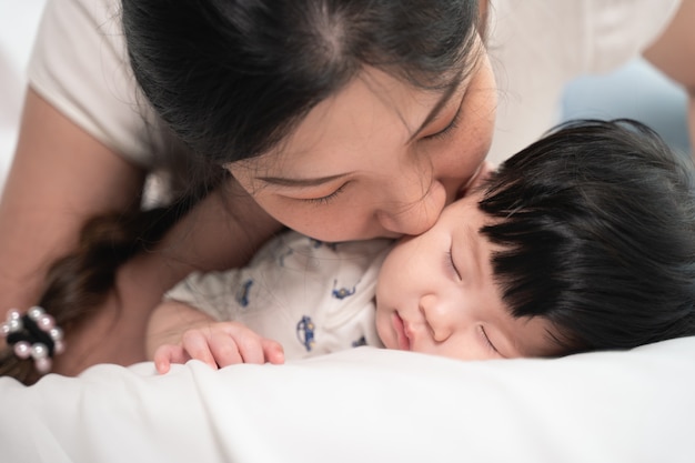 부드럽게 키스하고 사랑으로 침대에서 자고있는 아기를 만지고 키스하는 아시아 어머니는 행복을 느낍니다.