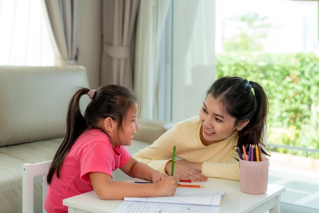 Asian mother playing with her daughter drawing together with color pencils at table in living room at home.