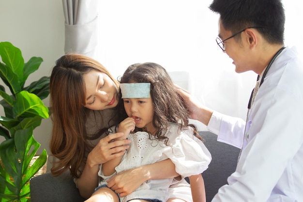 アジア人の母親が病気の若い娘を連れて病院の医者に診てもらい、医者が女の子の額に冷たい熱ゲルを貼り付けた女の子がインフルエンザウイルスで病気になった若い女の子の患者が病気になった