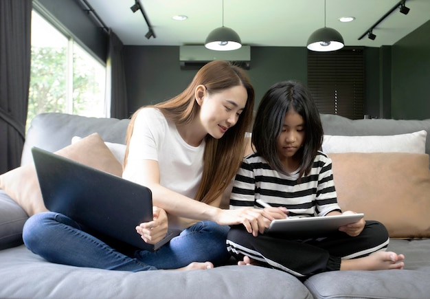 asian mother teaches her child in an online class with tablet and working on a laptop at home