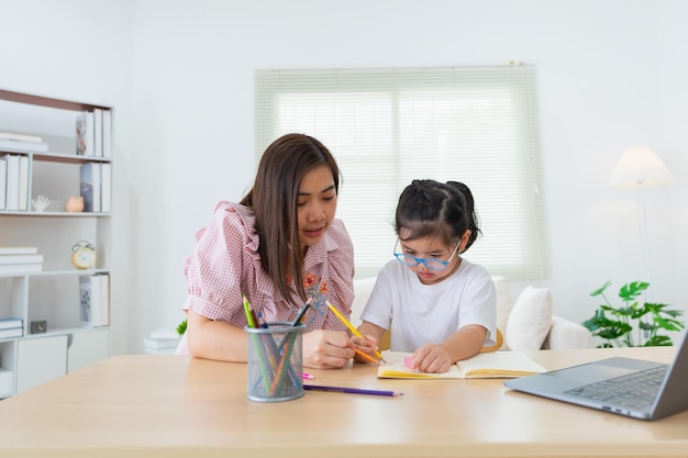 Asian mother teaching baby girl wearing eyeglasses doing home using laptop and write notes in notebook to study online on wood table in living room at home Education learning back to school concept