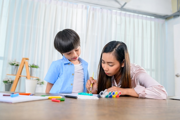 Asian mother work home together with son. Mom and kid drawing picture and color painting art. Woman lifestyle and family activity.