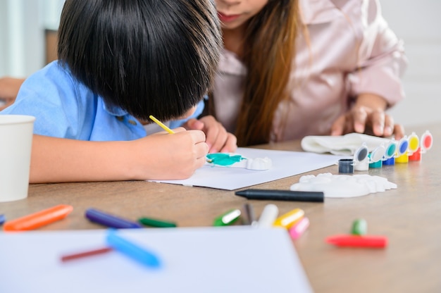 アジア人の母親は息子と一緒に家で働きます。ママと子供が絵とカラーペインティングアートを描きます。女性のライフスタイルと家族の活動。