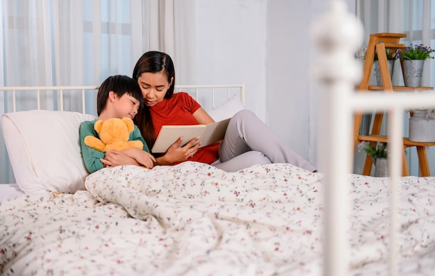 Asian mother work home together with son. Mom and kid reading fairy tale before sleep on the bed. Woman lifestyle and family activity.