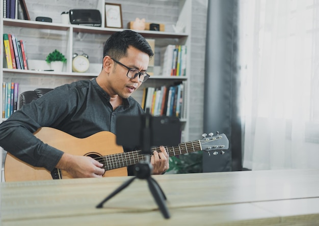 Asian music teacher man broadcasting live guitar lessons to students online using mobile Asian male musician playing guitar singing live video conferencing at home Live concert music from anywhere