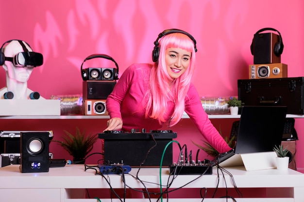 Asian musician standing at dj table wearing headset while playing techno music at professional mixer console in club at night time. Woman with pink hair having fun while enjoying performing song
