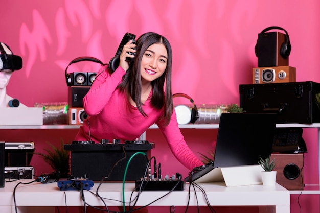 Asian musician wearing headphones while performing electronic music using professional turntables in studio over pink background. Dj playing stereo sounds with electronics and microphone