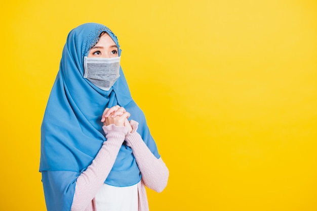 Asian Muslim Arab, Portrait of beautiful young woman Islam religious wear veil hijab and face mask protect quarantines disease coronavirus raising hand praying Eid al Fitr isolated yellow background