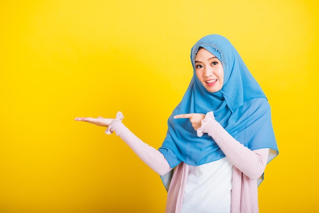 Asian Muslim Arab, Portrait of happy beautiful young woman Islam religious wear veil hijab funny smile she positive expression pointing finger side sideways to space isolated yellow background