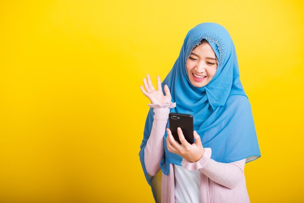 Asian muslim arab, portrait of happy beautiful young woman islam religious wear veil hijab funny smile she selfie or video call mobile smart phone raise hand say hello isolated on yellow background