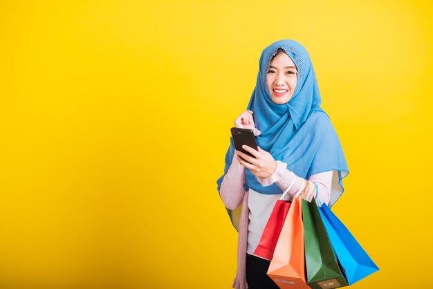 Asian muslim arab, portrait of happy beautiful young woman islam religious wear veil hijab funny smile she using smartphone on hand and hold shopping bags, studio shot isolated on yellow background
