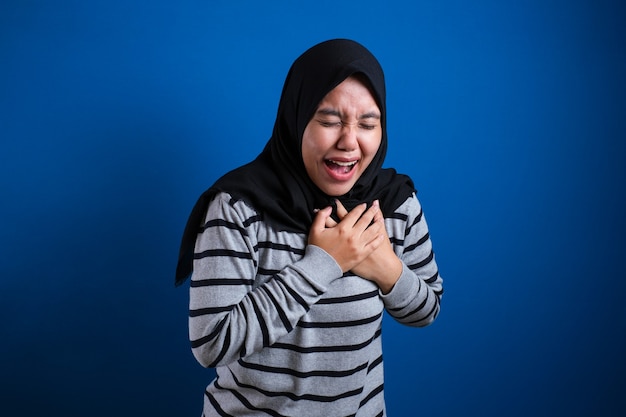 彼女の胸の痛みを感じているアジアのイスラム教徒の大学生の女の子、左胸を保持しているジェスチャー。青い背景
