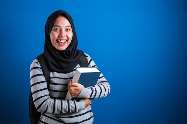 asian muslim college student girl wearing hijab smiling friendly holding big book