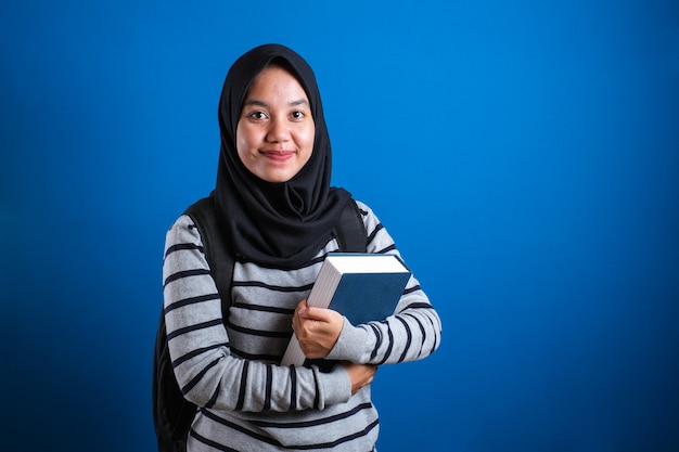 asian muslim college student girl wearing hijab smiling friendly holding big book