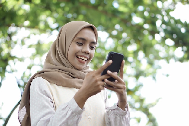 Asian Muslim girl in casual clothes is using a smartphone and smiling while standing outdoor area