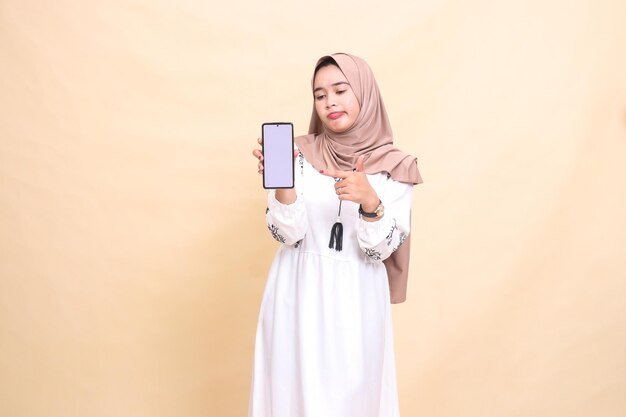 An Asian Muslim girl wearing a hijab frowns candidly showing a cellphone gadget screen pointing to c