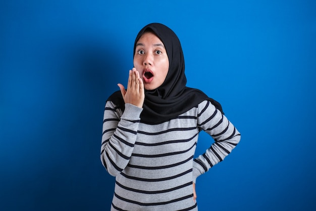 Asian muslim girl wearing hijab shows surprised or shocked expression with open mouth, close up facial expression over blue background