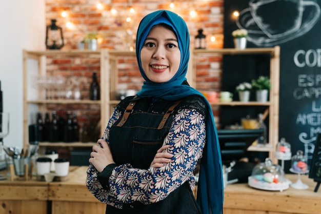 카페 바에서 웨이트리스로 일하는 히잡을 쓴 아시아 이슬람 소녀. 젊은 우아한 이슬람 여성이 팔짱을 끼고 커피숍에서 소규모 창업 사업을 시작했습니다. 아랍어 여자 직원 얼굴 카메라 미소 매력적인