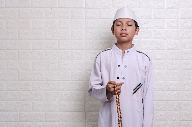 Asian muslim kid wearing white rob and skullcap holding prayer beads, closing eyes making dua