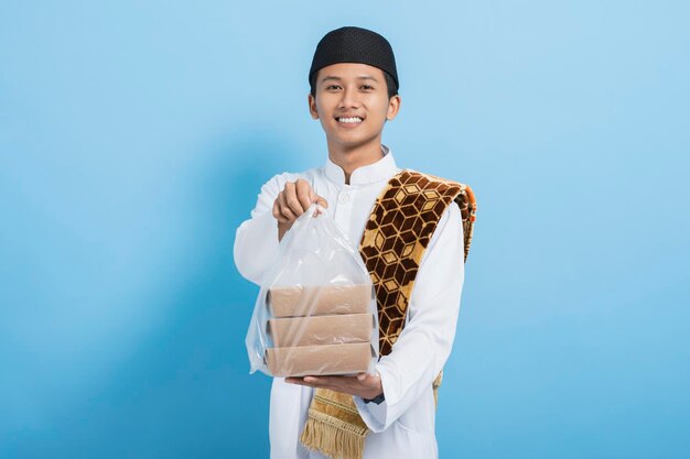 Asian muslim man carrying praying mat giving food for iftar at ramadan isolated on blue background