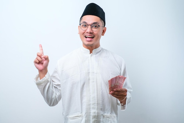 Asian muslim man holding rupiah paper money while pointing his finger up and smiling isolated on white background