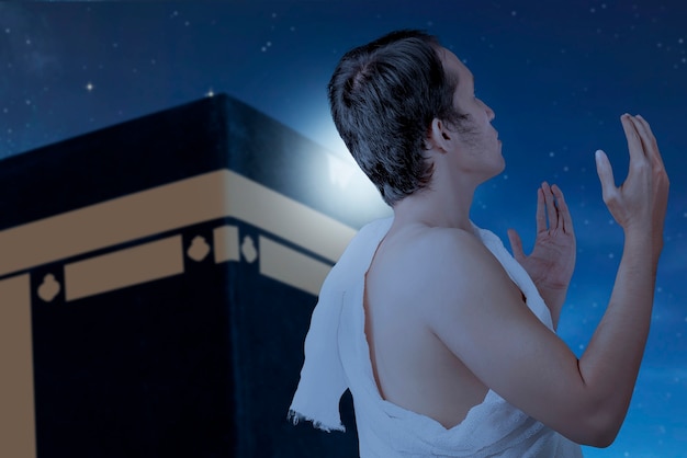 Asian Muslim man in ihram clothes standing and praying with Kaaba and night scene background