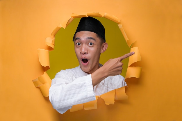 Asian Muslim man poses through torn yellow paper hole with shocked face, wearing muslim cloth with skull cap, pointing a copy space to presenting something.