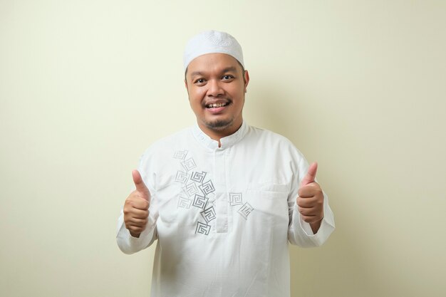 Asian Muslim man smiled looking at the camera, with victory gesture. Isolated on ivory background