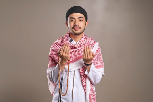 Foto uomo musulmano asiatico che indossa kufiya e cappello da preghiera mentre prega mentre alza le mani