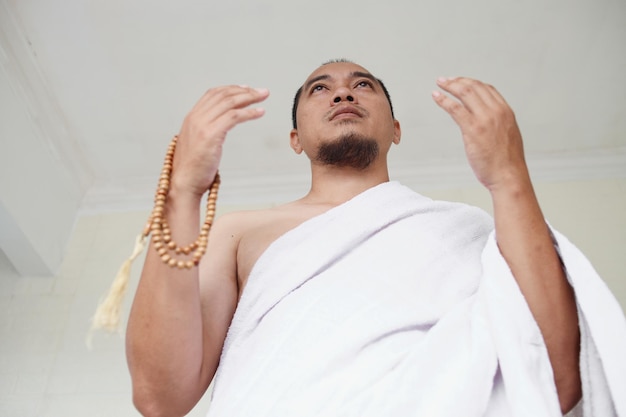 Asian muslim man wearing white ihram clothes and praying while holding prayer beads