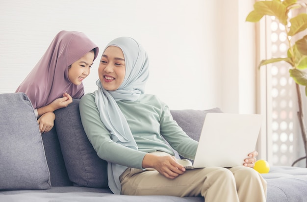 Asian muslim mother and daughter smiling when using laptop together, browsing internet or watching streaming movie