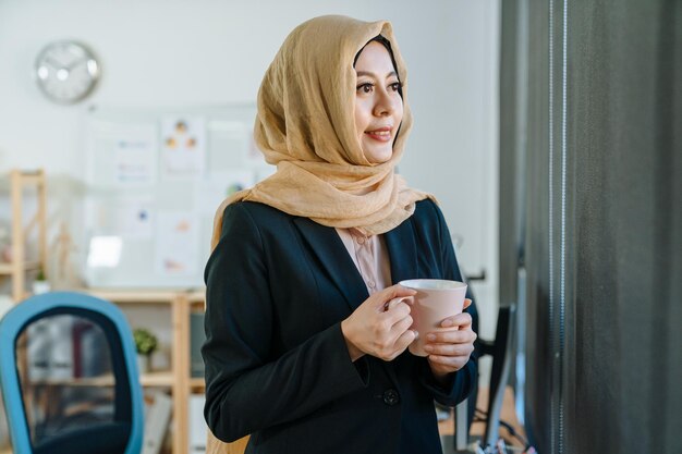 Asian muslim woman enjoying cup of tea in coffee break time in\
modern cozy office. islam female employee in head scarf and suit\
stand by window look city view with sunshine on face charming\
smiling