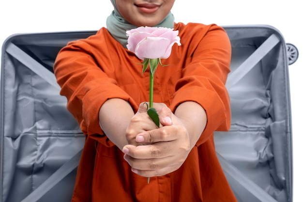 Asian Muslim woman in a headscarf sitting in a suitcase while holding a pink rose flower