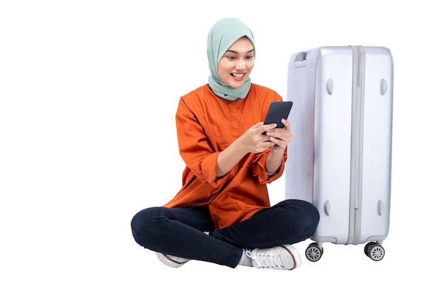 Asian Muslim woman in a headscarf sitting with a suitcase while using a mobile phone